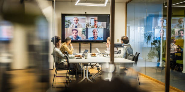 Business people attending a hybrid meeting with some people in a conference room and others attending via video conferencing on a large screen.